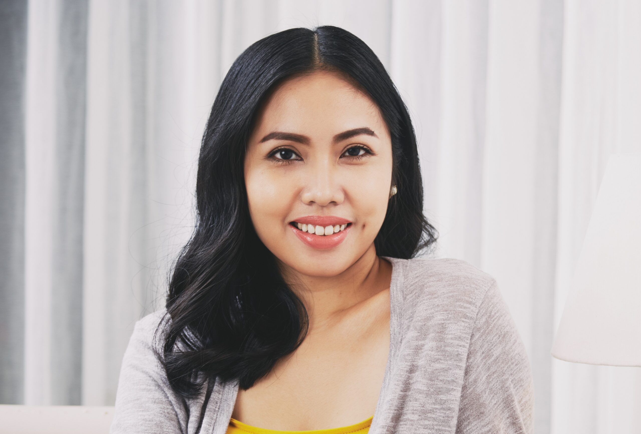 Positive Filipino female smiling and looking at camera while sitting near lamp in stylish living room